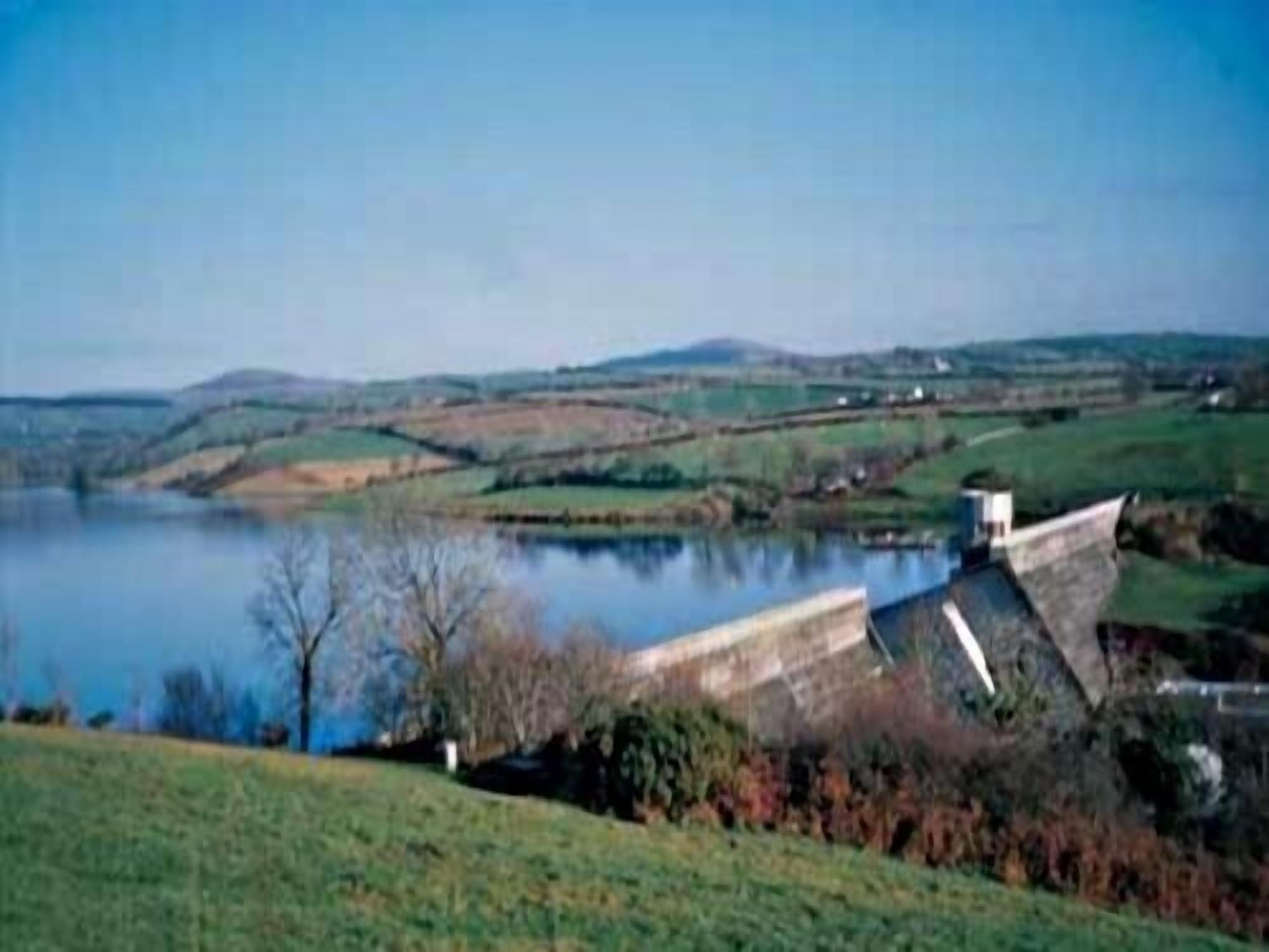 Un barrage surmonté d’un petit bâtiment, entouré de champs verdoyants et bordé par une étendue d’eau.