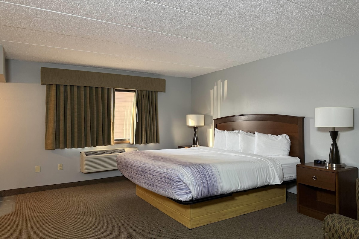 A hotel room with a bed, bedside lamps, a nightstand, a window with curtains, and an air conditioning unit.