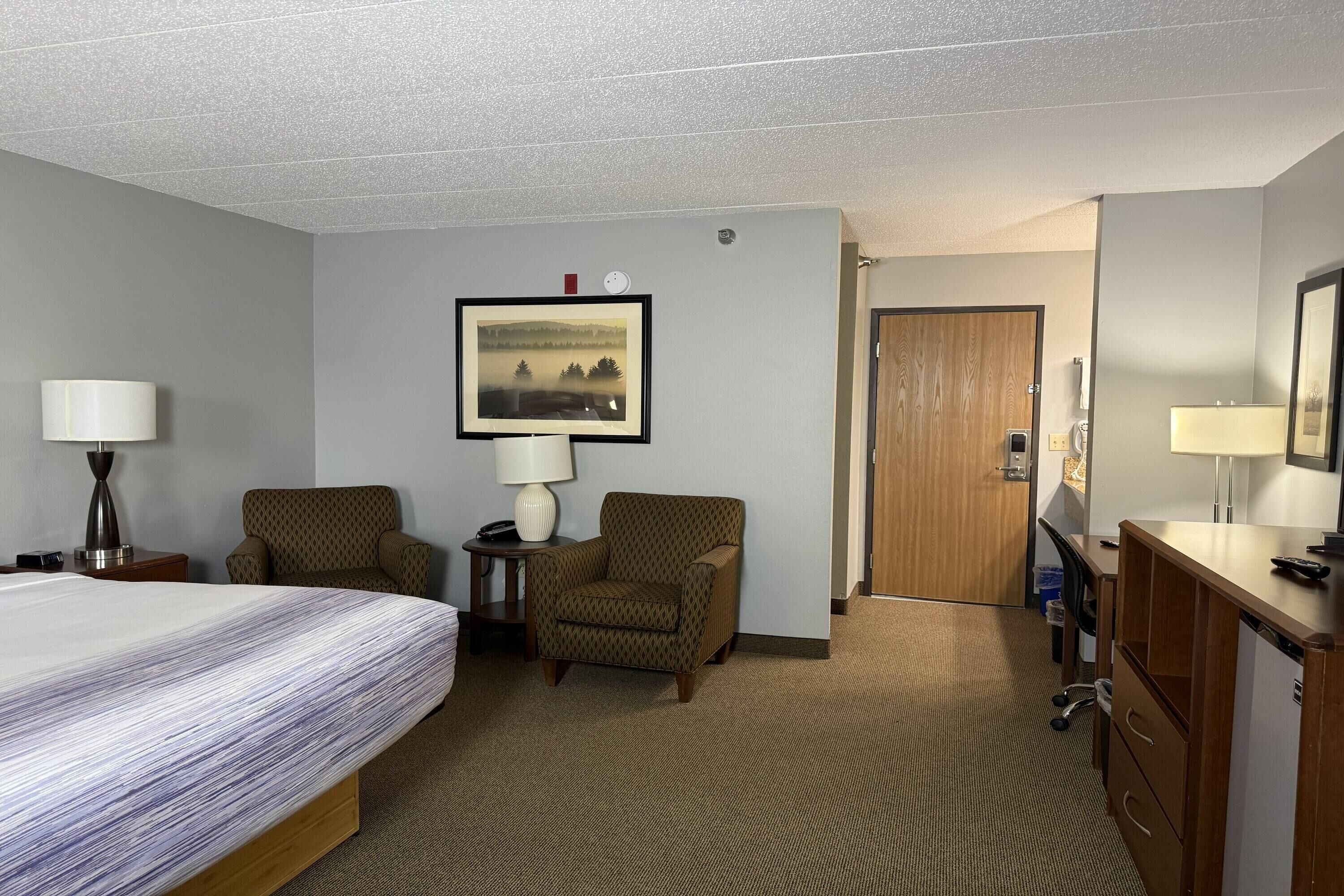 Habitación de hotel con una cama, dos sillones, un escritorio, un aparador, una lámpara, un cuadro en la pared y una puerta que da a otra habitación.