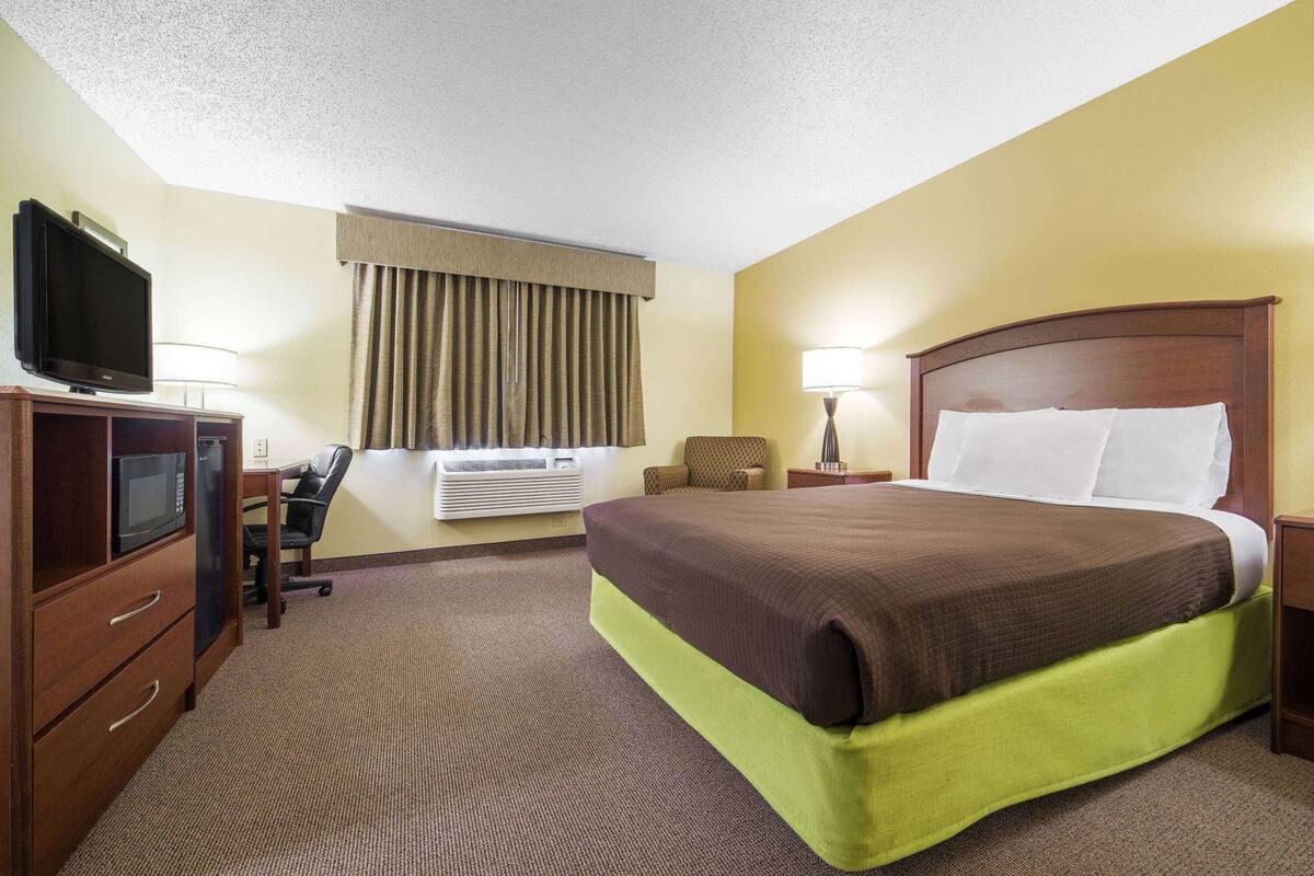 A hotel room with a bed, a desk with a computer, a television, an air conditioning unit, and a window with curtains.