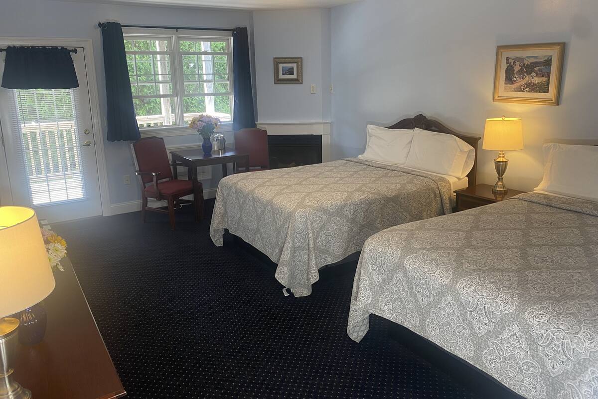 A hotel room with two beds, a desk, a chair, a fireplace, and a window with curtains.