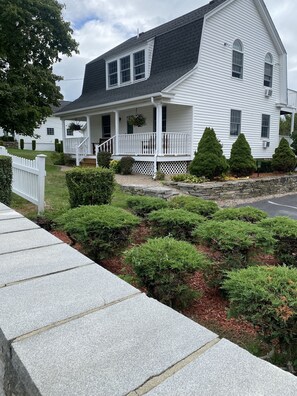 A white two-story house with a porch, a stone wall, and well-maintained bushes.