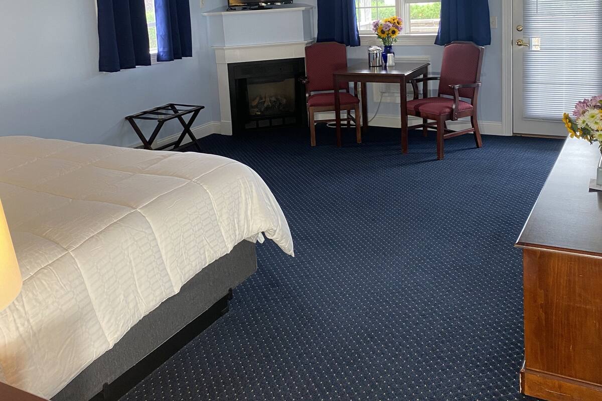A hotel room with a bed, a TV, a fireplace, a dining table with chairs, and a window with curtains.