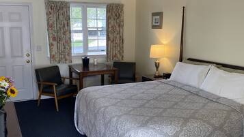 Habitación de hotel con una cama grande, un escritorio con dos sillas, una ventana con cortinas florales, una lámpara de noche y un cuadro en la pared.
