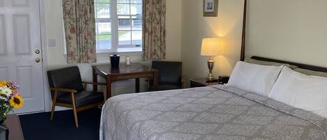 A hotel room with a large bed, a desk with two chairs, a window with floral curtains, a bedside lamp, and a framed picture on the wall.