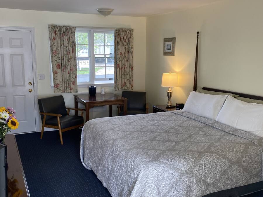 A hotel room with a large bed, a desk with two chairs, a window with floral curtains, a bedside lamp, and a framed picture on the wall.