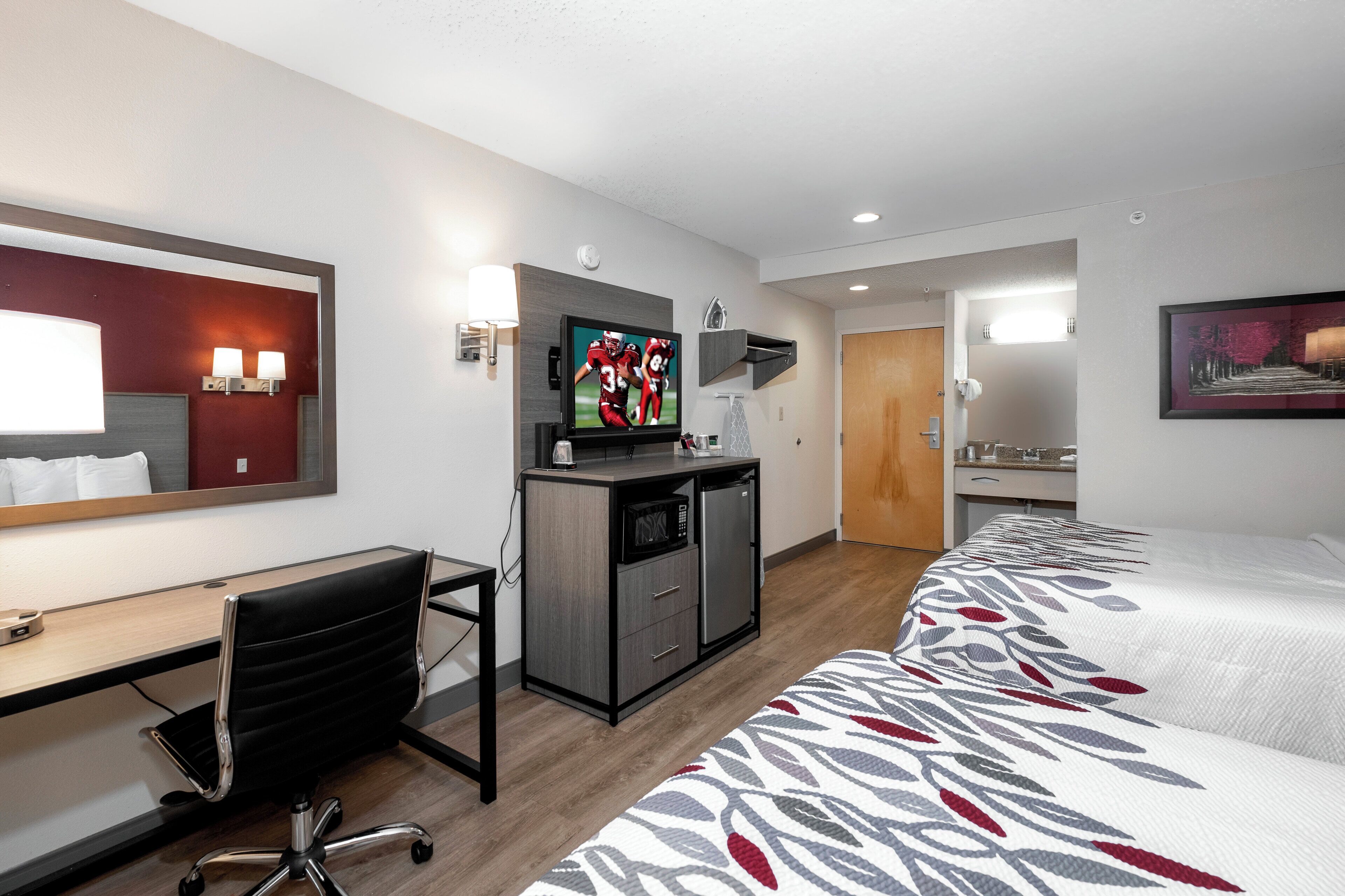 A hotel room with a desk, chair, television, minibar, and a bed with a patterned bedspread.
