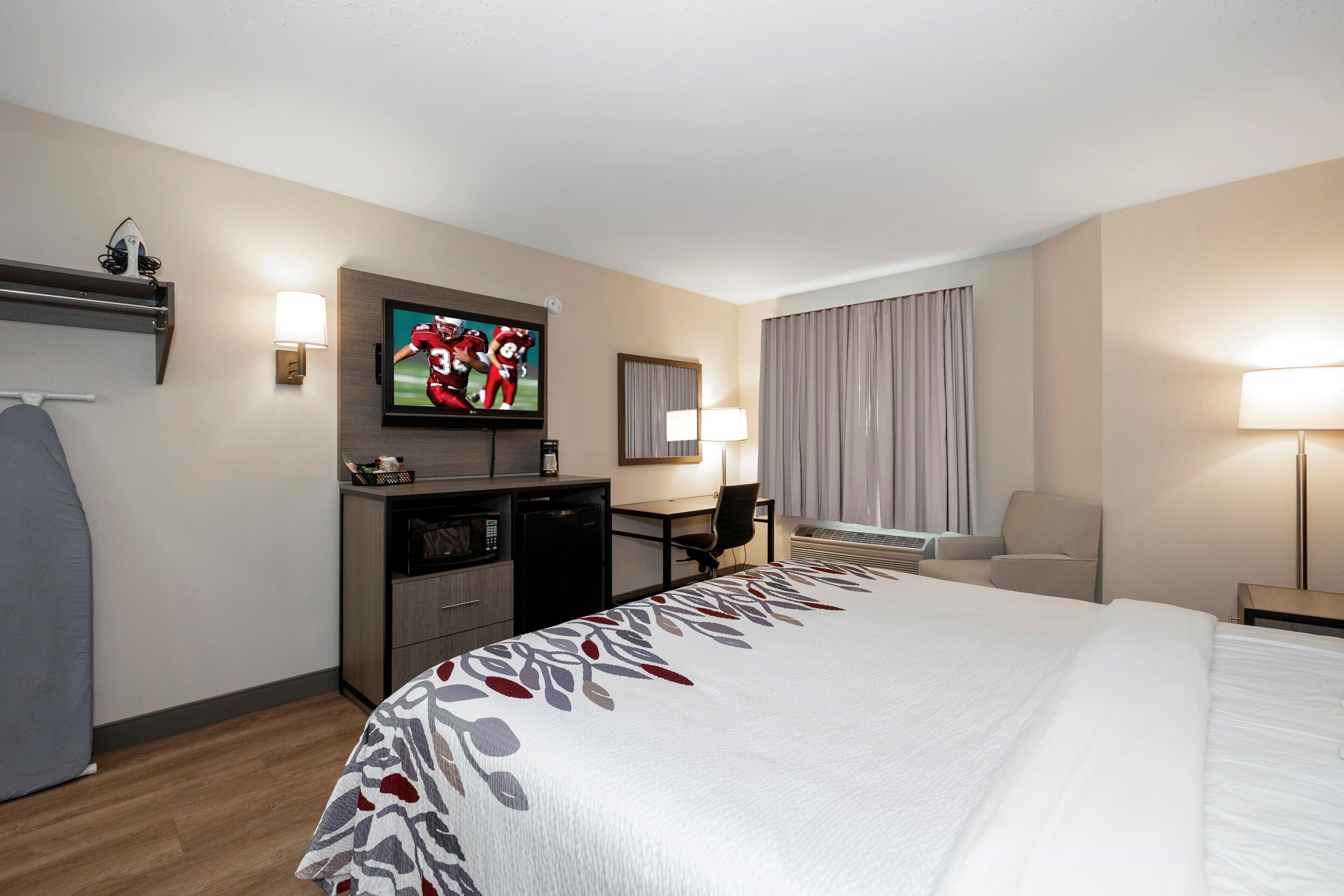 Ein Hotelzimmer mit einem großen Bett, einem Schreibtisch mit Computer, einem an der Wand montierten Fernseher und einem Fenster mit Vorhängen.