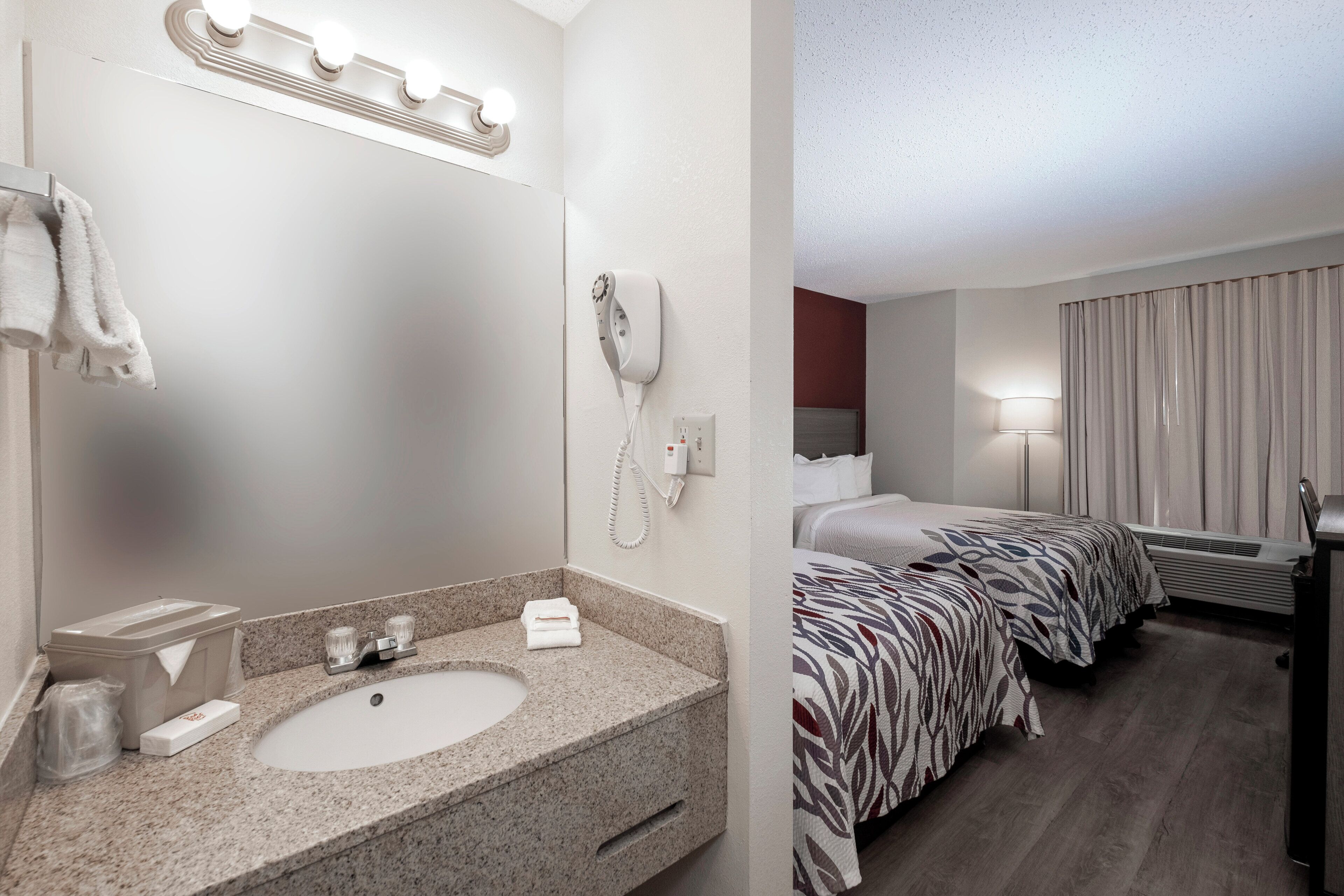 Een hotelkamer met een wastafel van graniet, een spiegel, een telefoon, een bed met een gestreept laken en een raam met gordijnen.