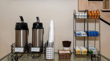 Estação de autoatendimento com dispensadores de café e água quente, copos de papel e diversos artigos de higiene pessoal nas prateleiras.