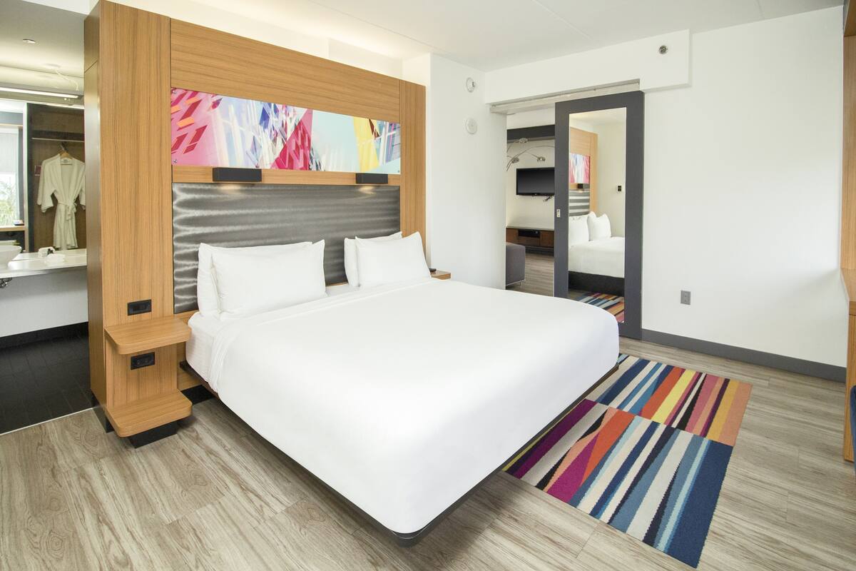 A modern hotel room with a large bed, wooden headboard, a colorful abstract painting, a mirror, and a striped rug.
