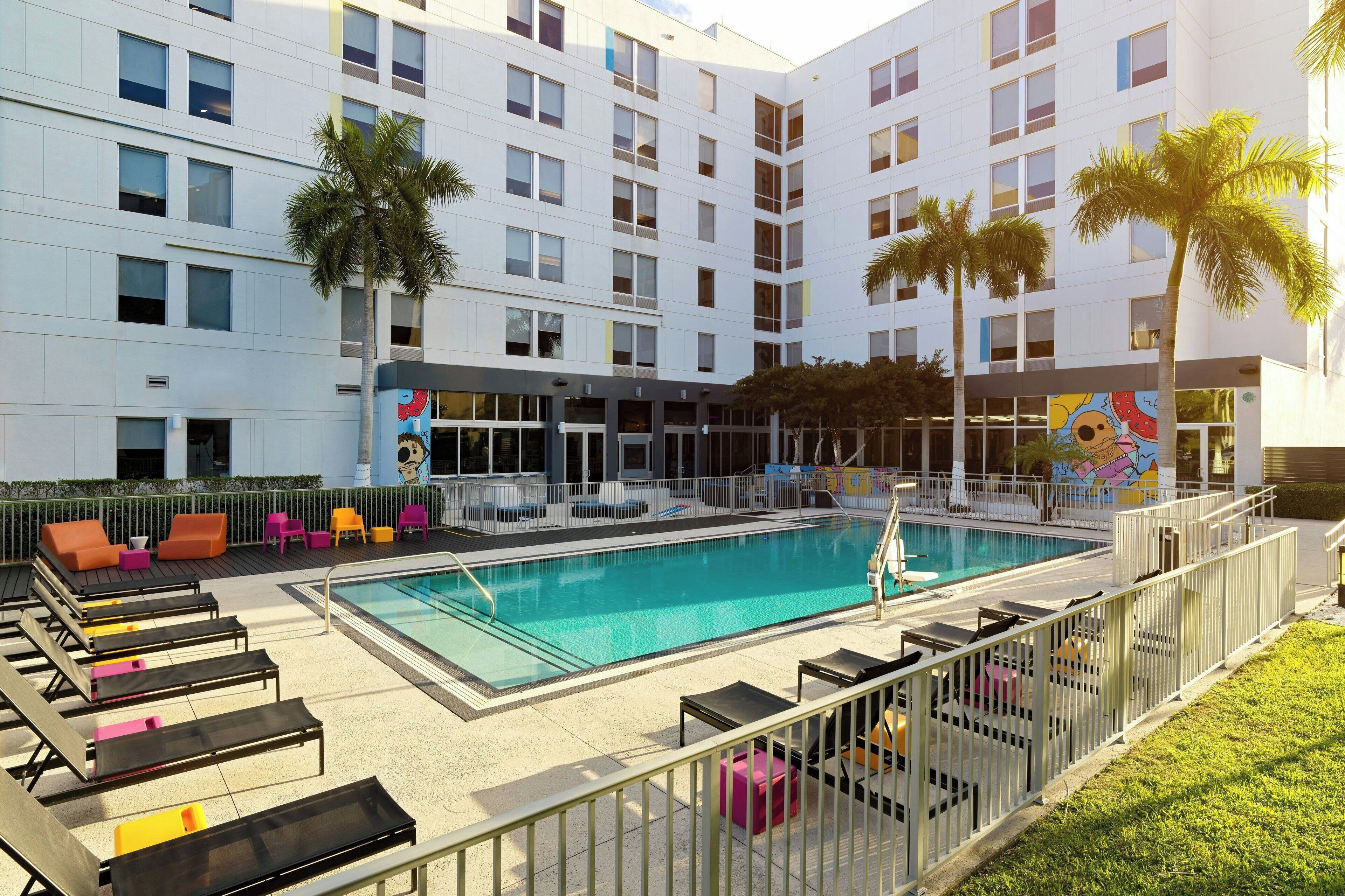A hotel pool area with lounge chairs, a swimming pool, and palm trees.