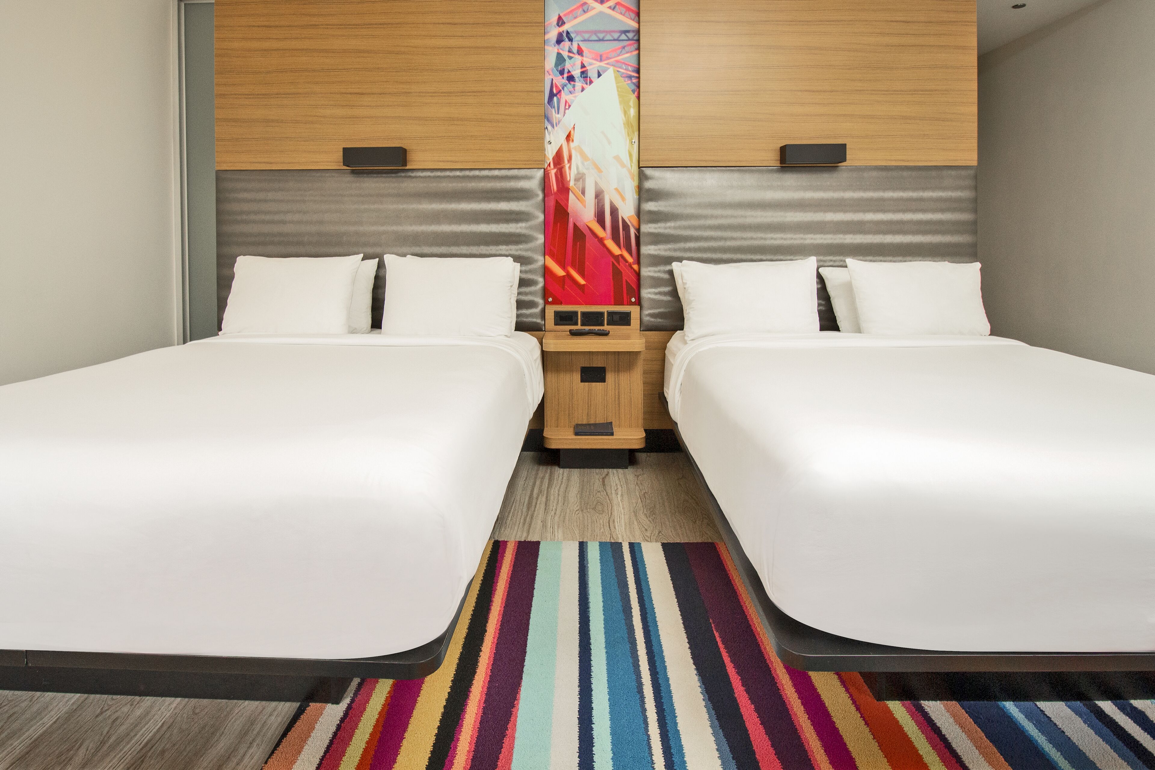 A hotel room with two beds, a colorful striped rug, and a wooden headboard with a decorative panel.