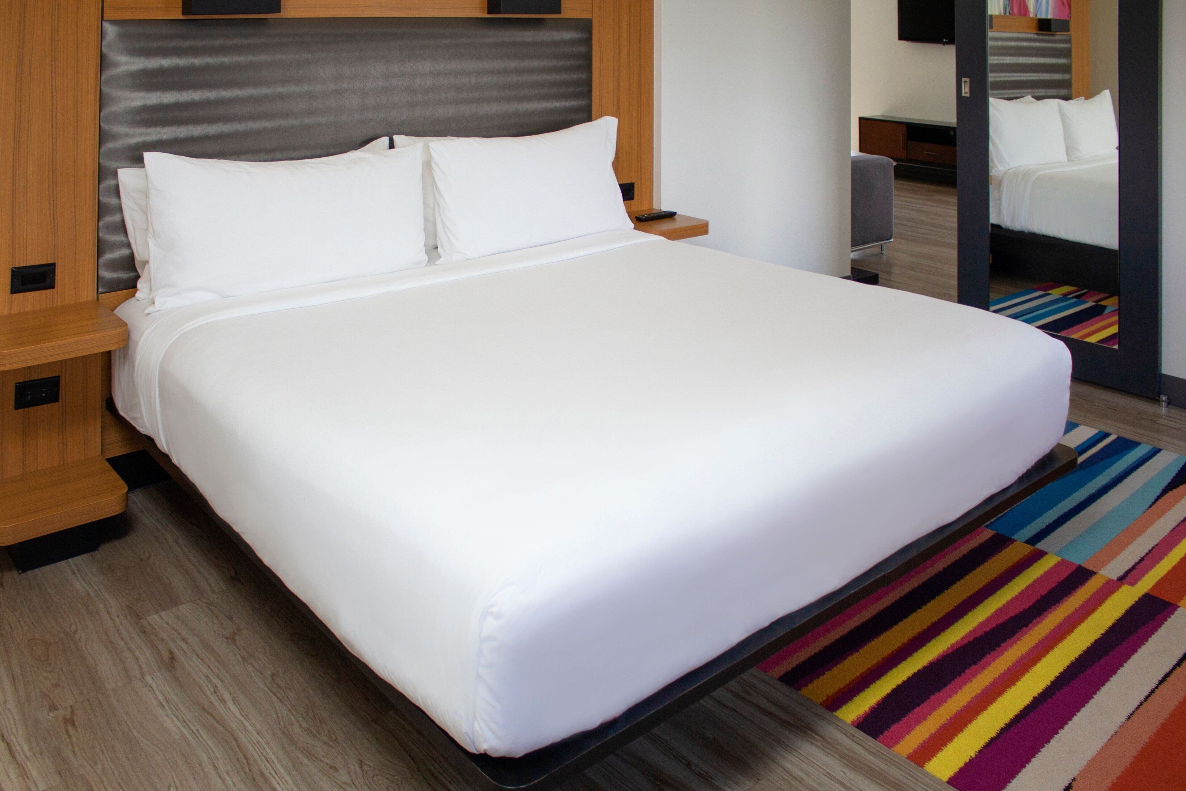 Un lit bien fait, avec du linge de lit blanc, une tête de lit en bois et un tapis rayé aux couleurs vives sur le sol.
