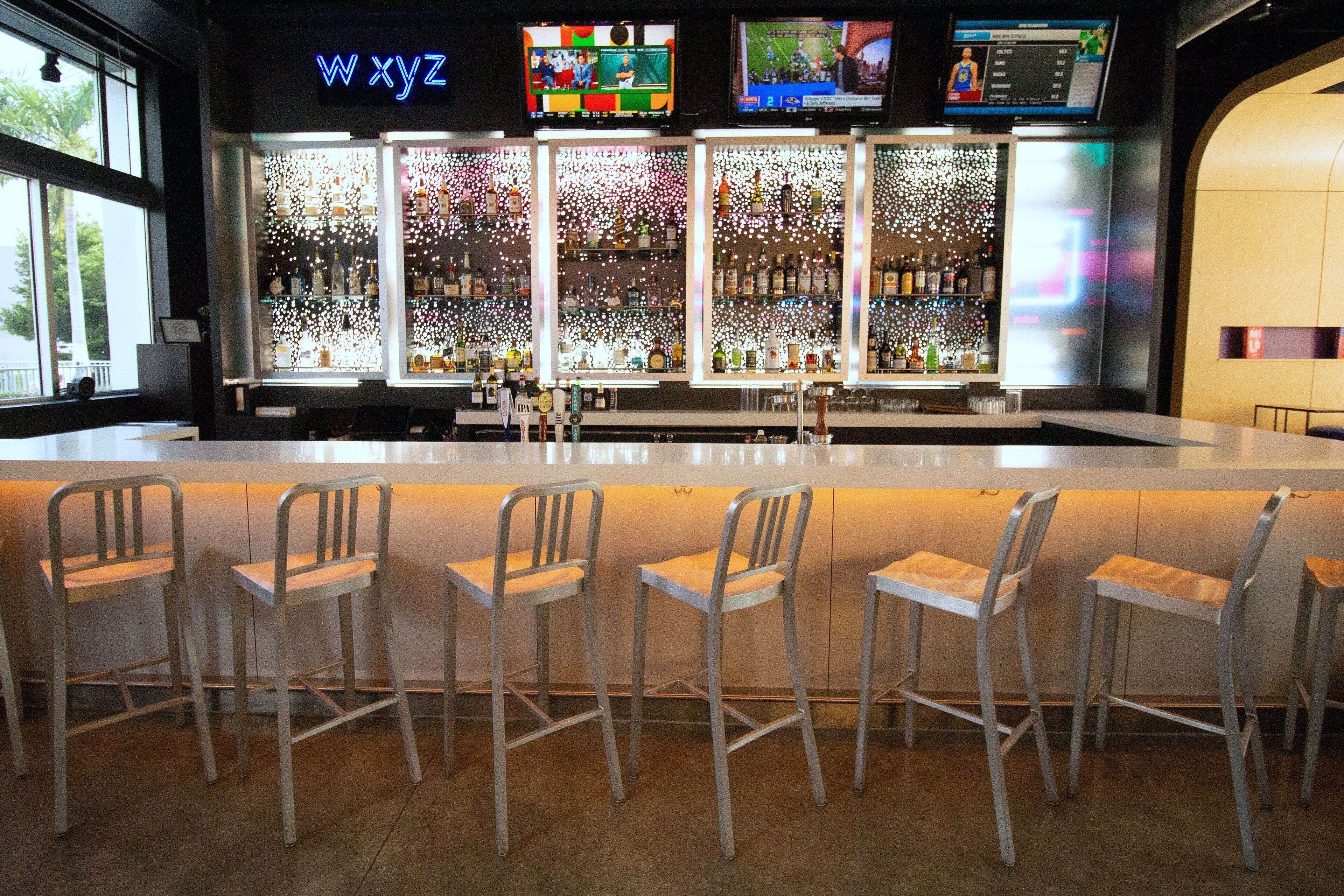 Zona de bar con un letrero de neón, varias pantallas y una amplia variedad de botellas.