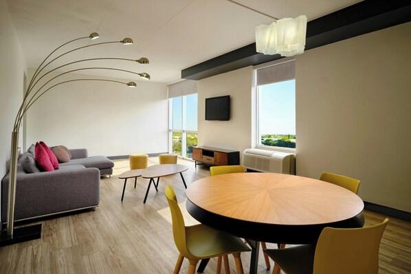 A modern living room with a sofa, a round wooden table, and yellow chairs. There is a TV mounted on the wall and a window providing a view outside.