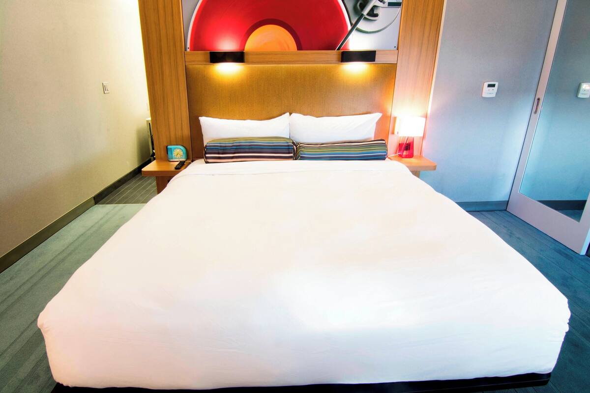 Habitación de hotel con una cama grande, cabecera de madera, mesitas de noche con lámparas y un adorno rojo en la pared.