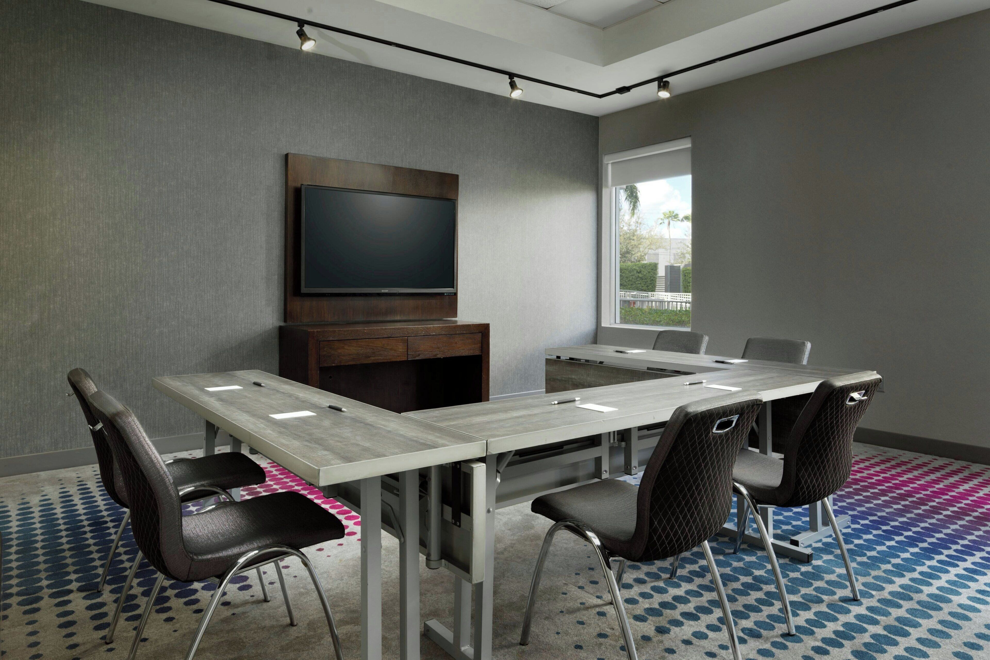A conference room with a long table, six chairs, a flat-screen TV, and a window with a view of buildings and trees.