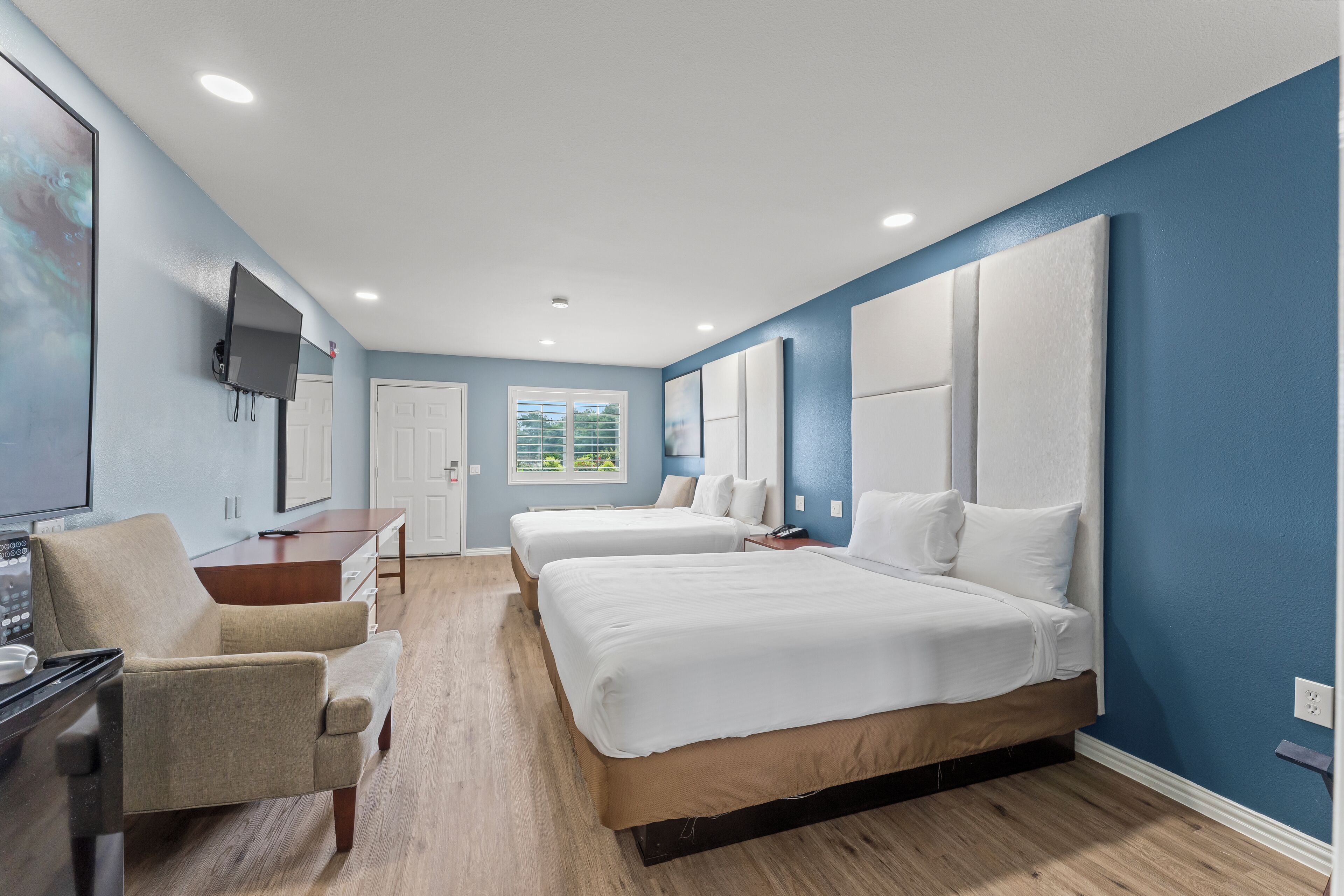 A hotel room with two beds, a desk, a chair, a TV, and a window with a view of greenery.