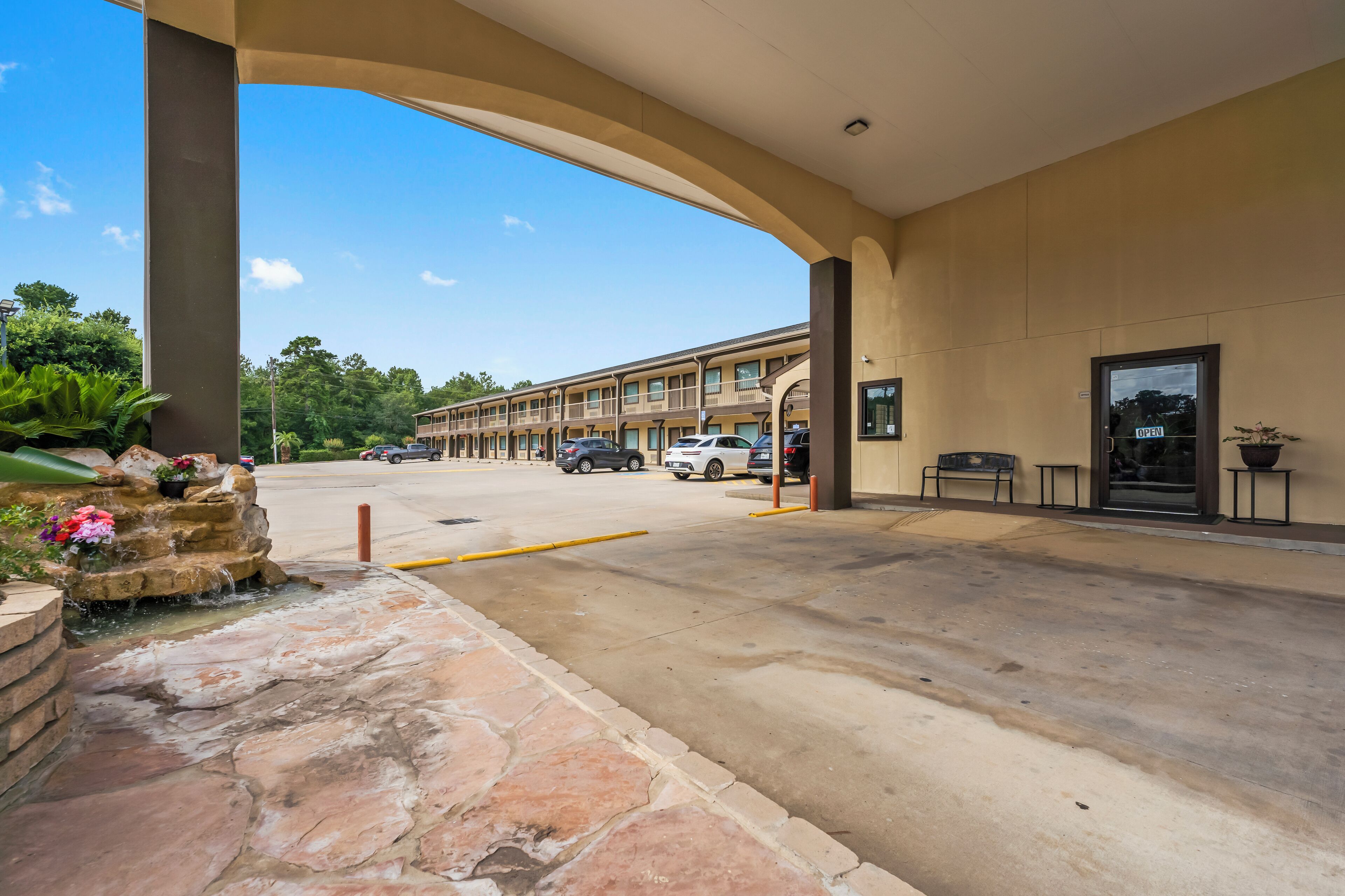 A hotel entrance with a covered walkway, a stone planter with flowers, and a row of parked cars.