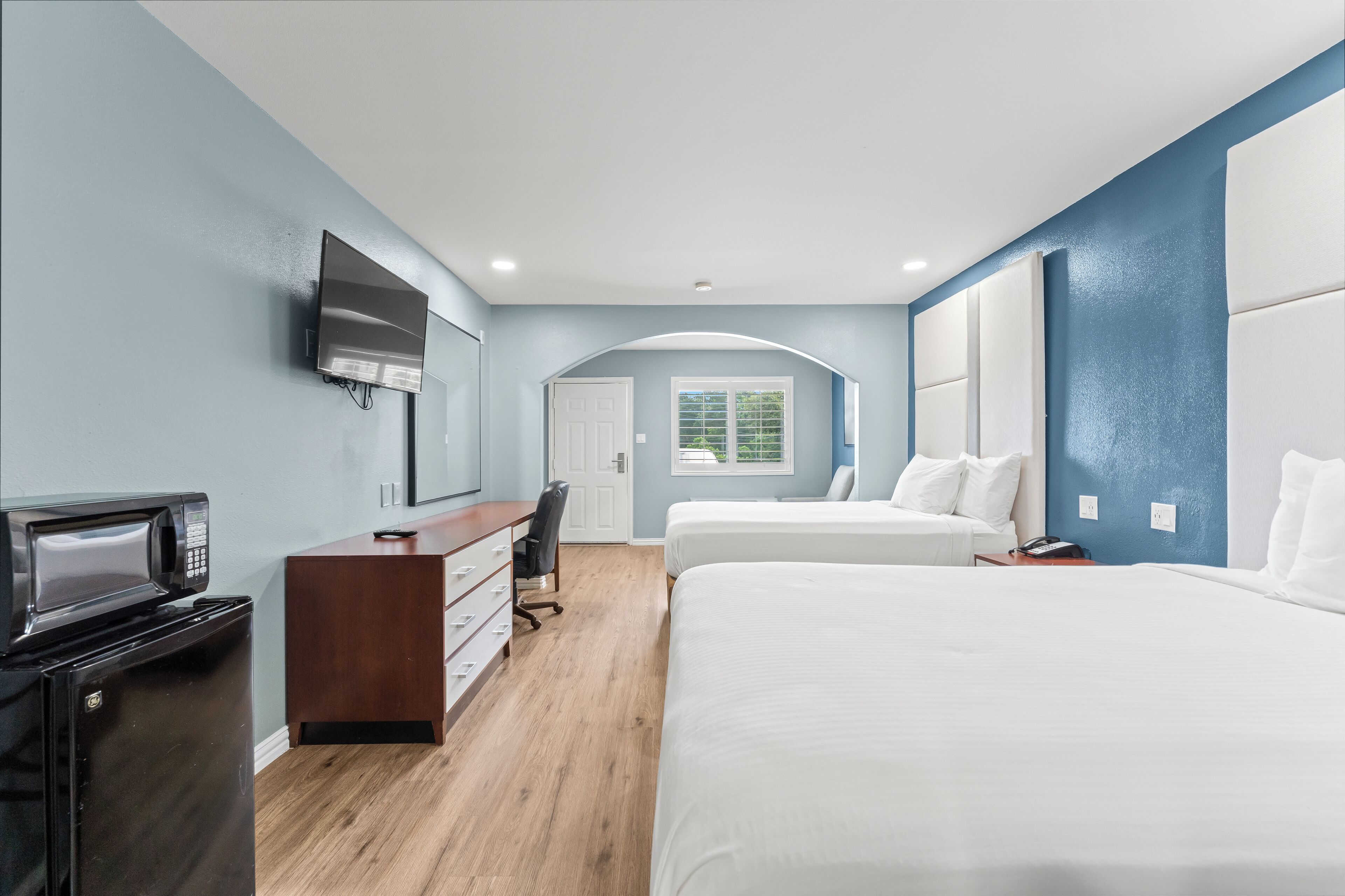 A modern hotel room with a flat-screen TV, a compact kitchenette, a desk, and a large bed.