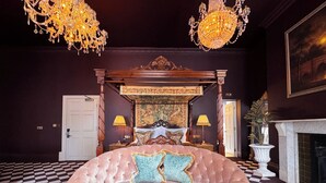 Een slaapkamer met een hemelbed, twee kroonluchters, een zwart-wit schakeleenvloer, een open haard en een groot schilderij aan de muur.