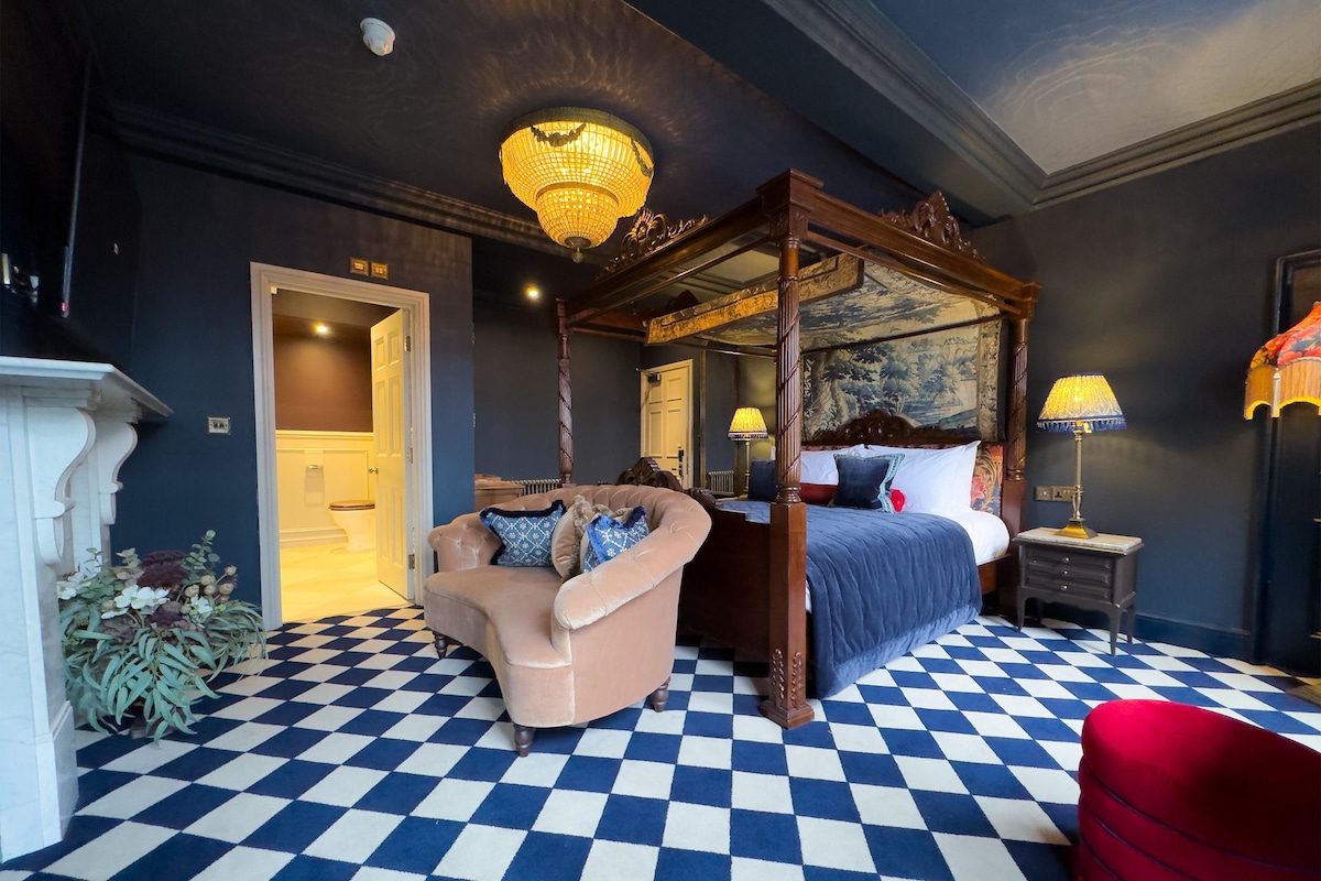 A room with a checkered floor, a four-poster bed, a chaise lounge, and a lamp on a nightstand.