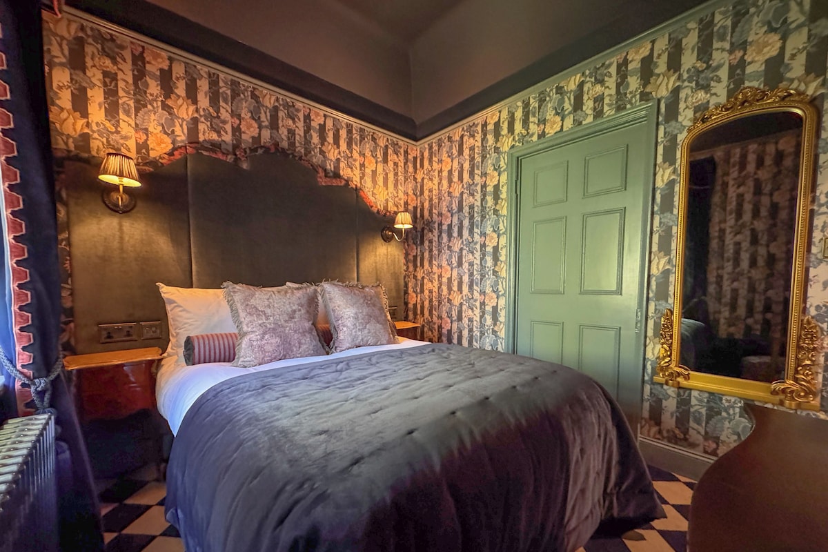 A bedroom with a large bed, a green door, a mirror with a gold frame, and patterned wallpaper.