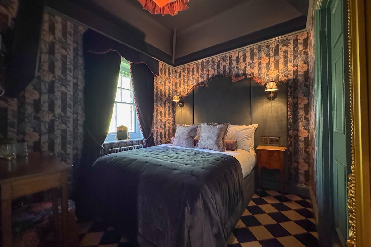 A bedroom with a checkered floor, a bed with multiple pillows, a nightstand, a window with curtains, and a wall with patterned wallpaper.