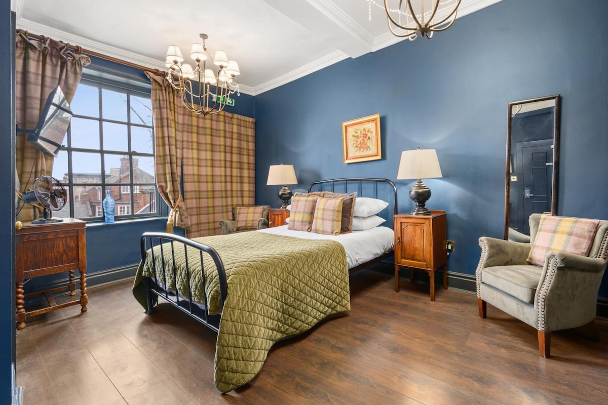 A bedroom with a bed, bedside tables, a chair, a window with curtains, a chandelier, and a framed picture on the wall.