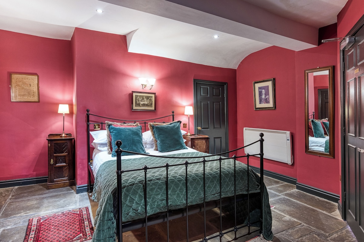 A bedroom with a four-poster bed, a red wall, a wooden nightstand, a bedside table with a lamp, and a door with a mirror.