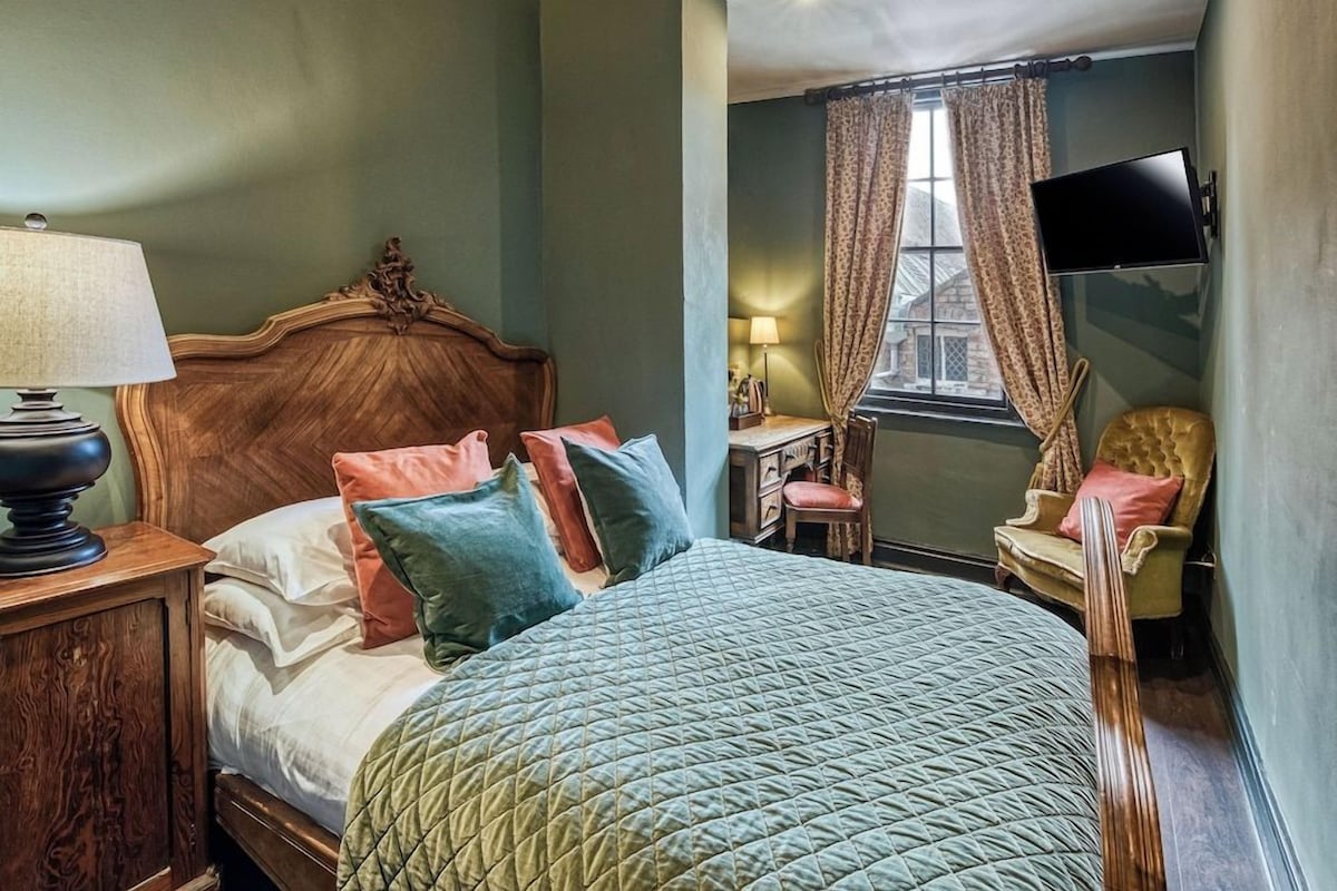 A bedroom with a wooden headboard, a bed with multiple pillows, a nightstand with a lamp, a chair, and a window with curtains.