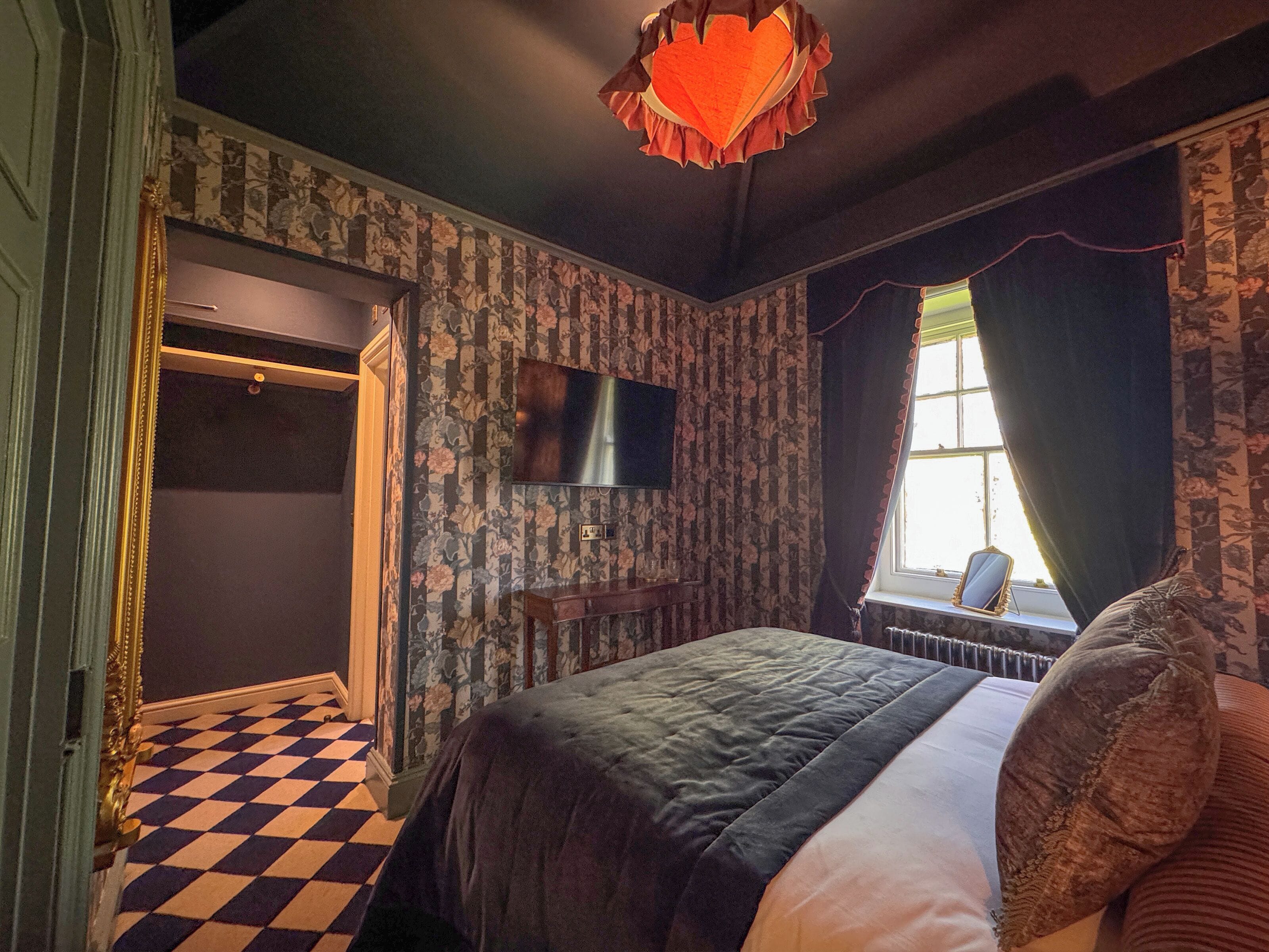 A bedroom with a checkered floor, a bed with a dark quilt, a television on the wall, and a window with curtains.