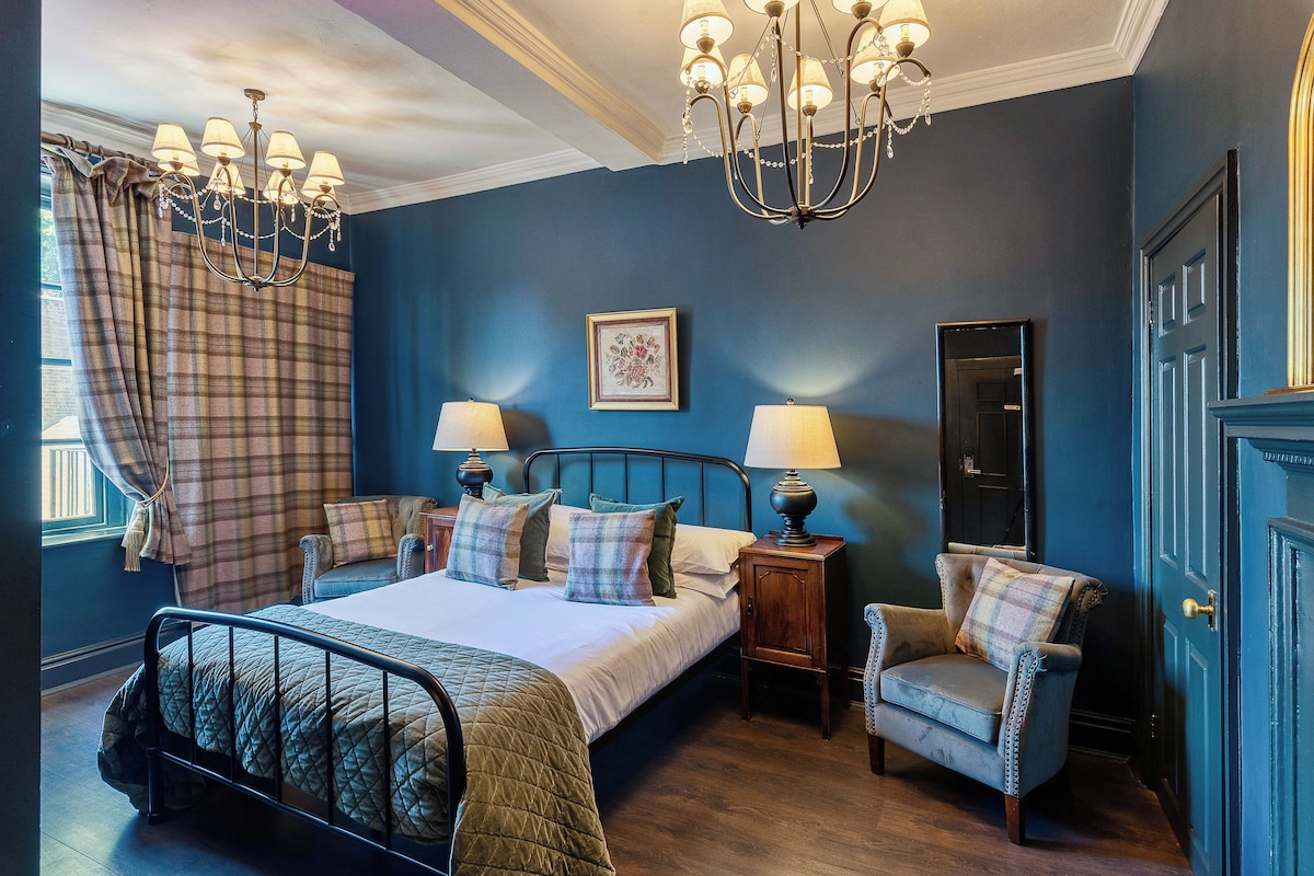A bedroom with a bed, two bedside tables, a chair, a chandelier, and a door.