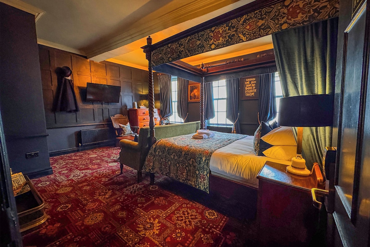 A four-poster bed with a canopy, a television mounted on the wall, a red patterned carpet, and a window with curtains.