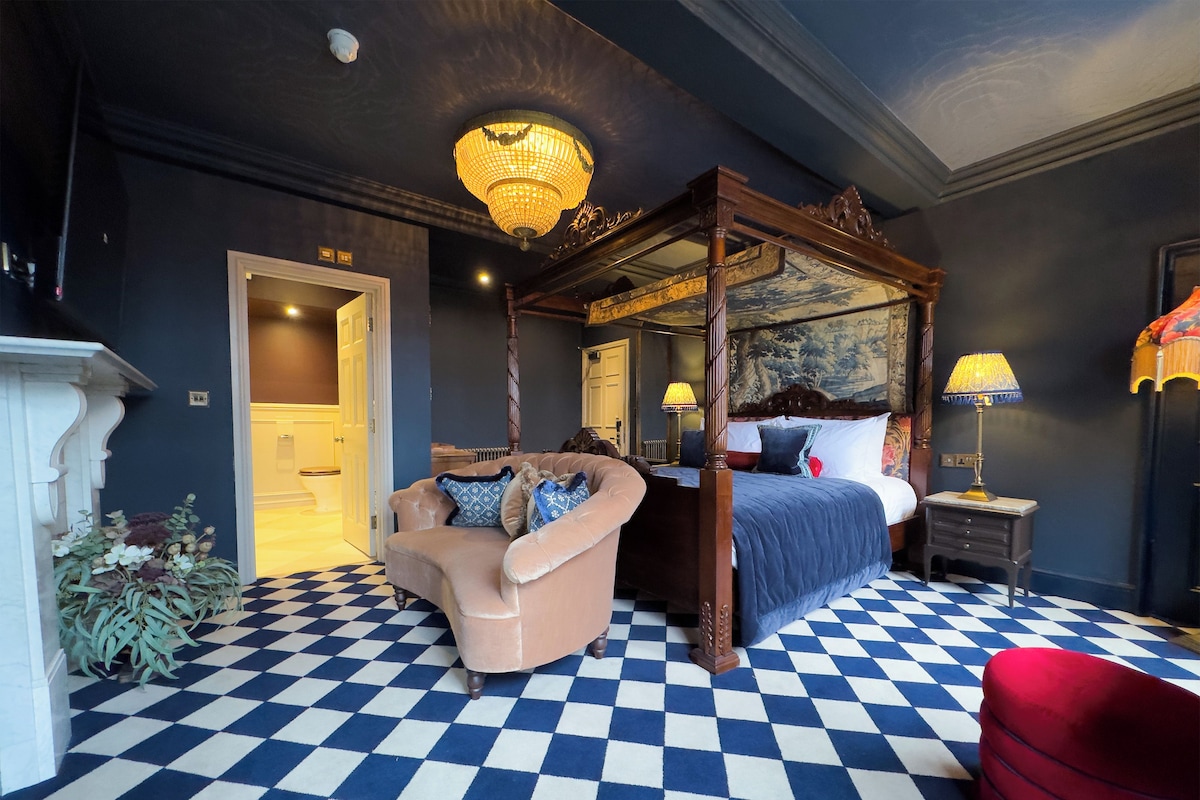 A room with a checkered floor, a four-poster bed, a chaise lounge, a lamp, and a painting on the wall.
