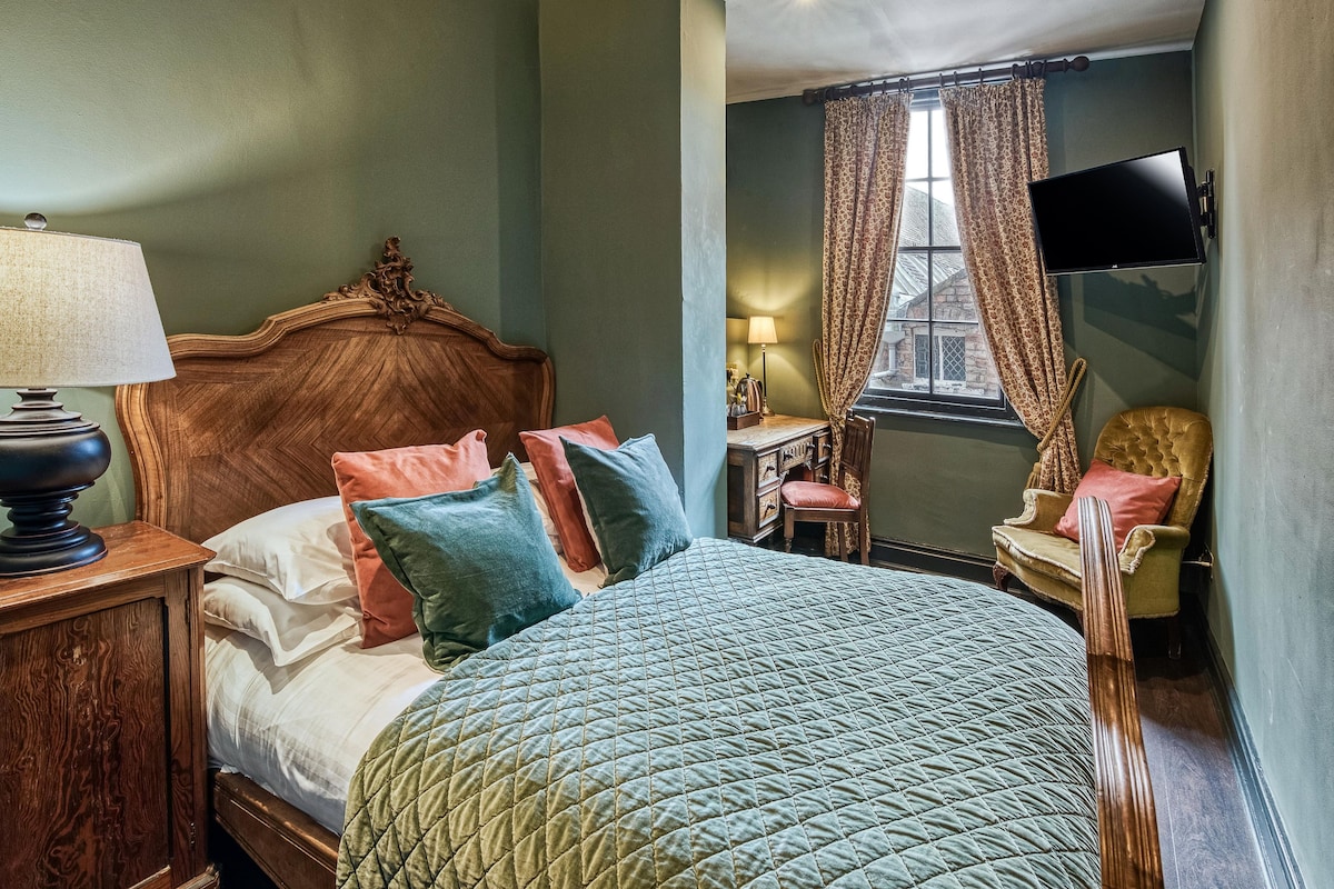A traditional bedroom with a wooden bed, multiple pillows, a nightstand, a desk with a lamp, a chair, and a television.
