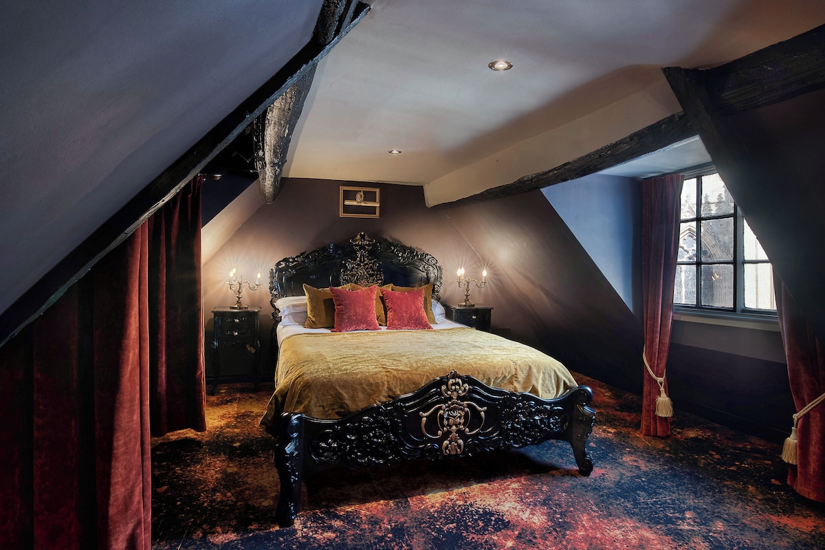 A bedroom with a high ceiling, a large bed with ornate headboard, and a window with curtains.