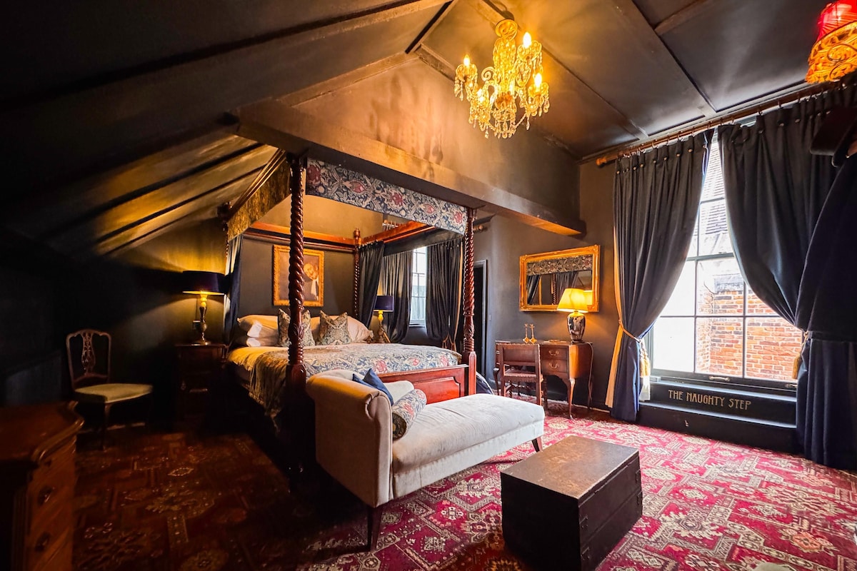 A four-poster bed with a canopy, a desk with a lamp, a chair, and a window with curtains.