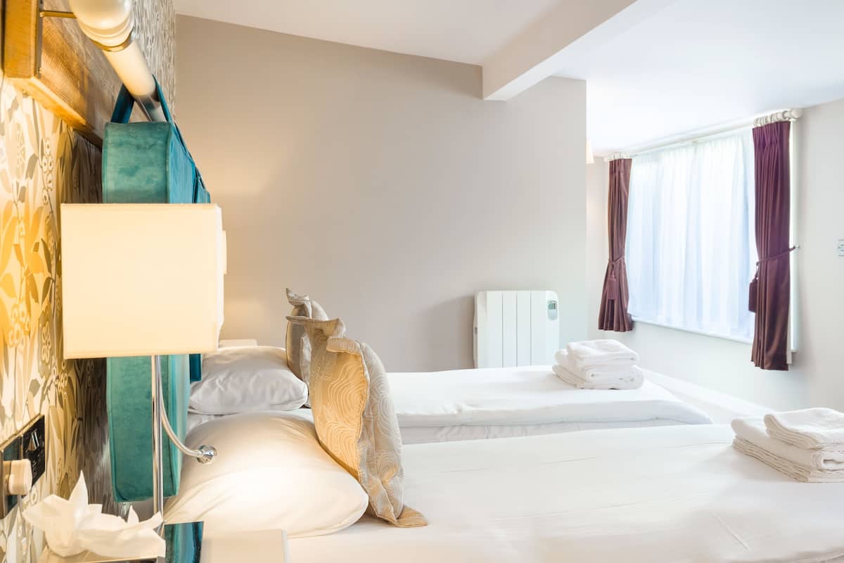 Una habitación de hotel con dos camas, una mesita de noche con lámpara, un radiador y una ventana con cortinas.