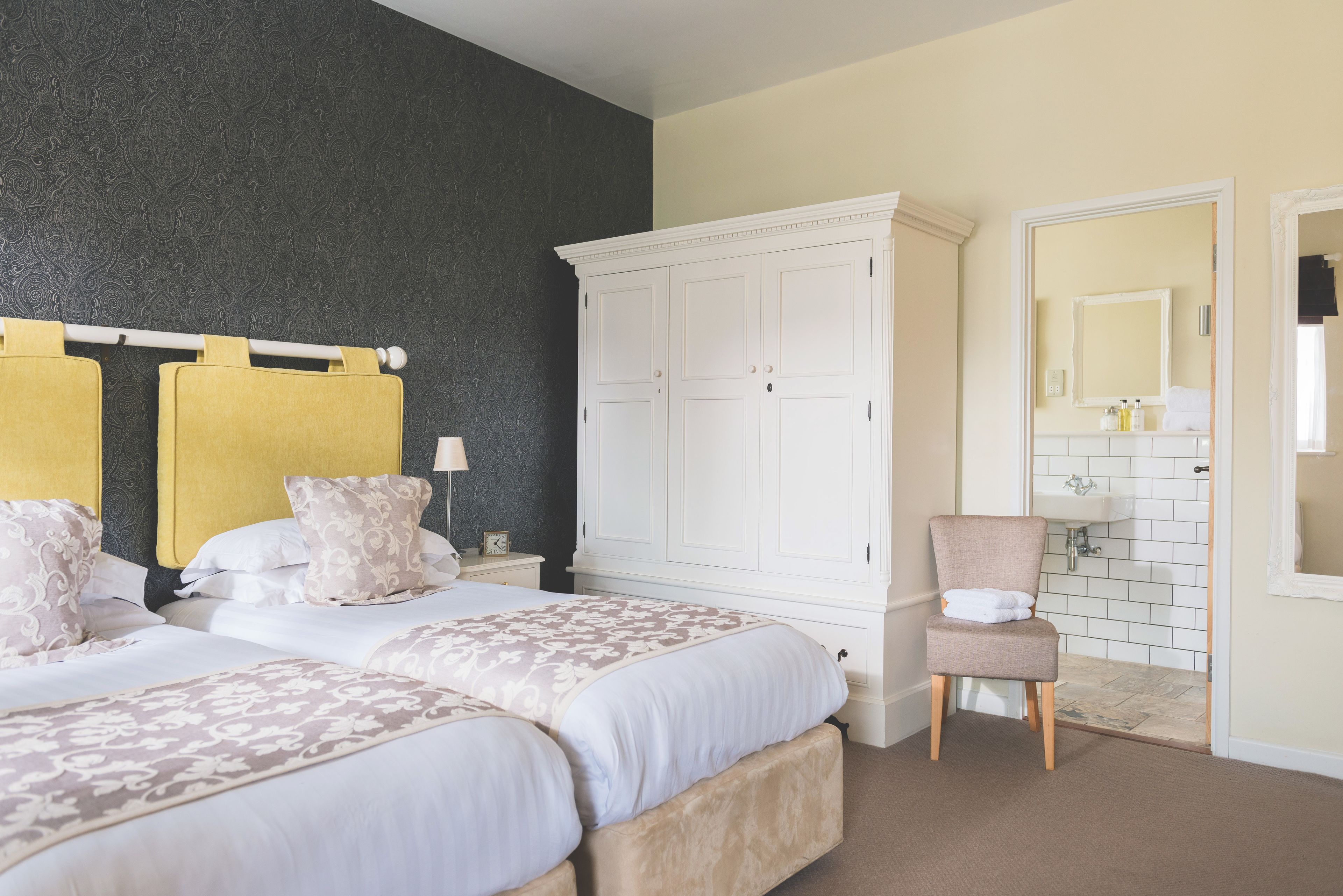 A bedroom with two beds, a large white wardrobe, a chair, and a bathroom visible through an open door.