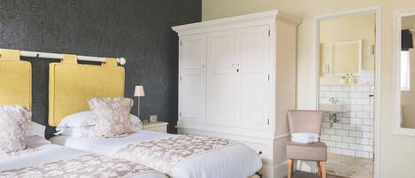 Un dormitorio con dos camas, un armario blanco grande, una silla y un baño visible a través de una puerta abierta.