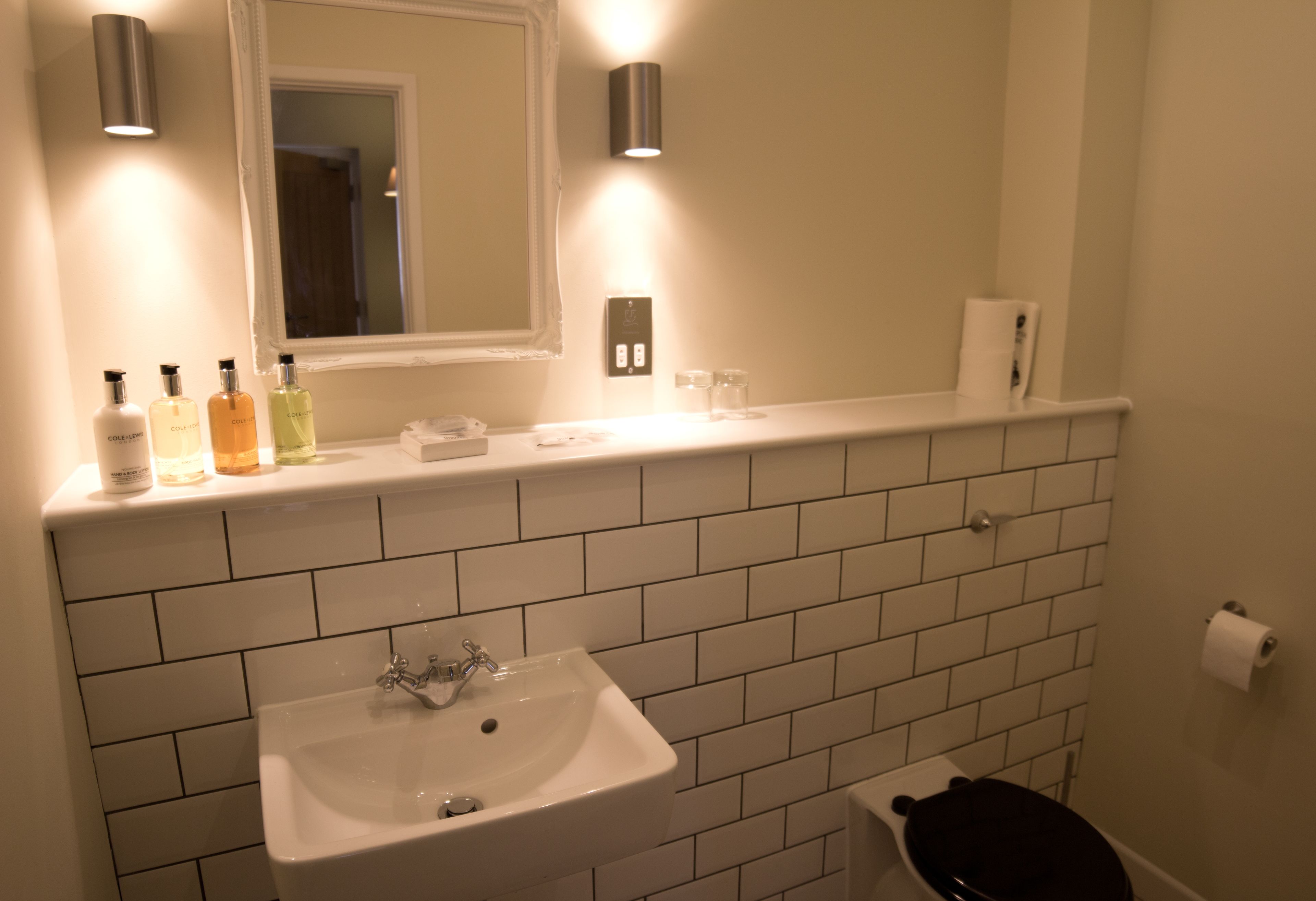 A bathroom with a white sink, mirror, and tiled walls. There are toiletries on the countertop and a toilet paper holder on the wall.