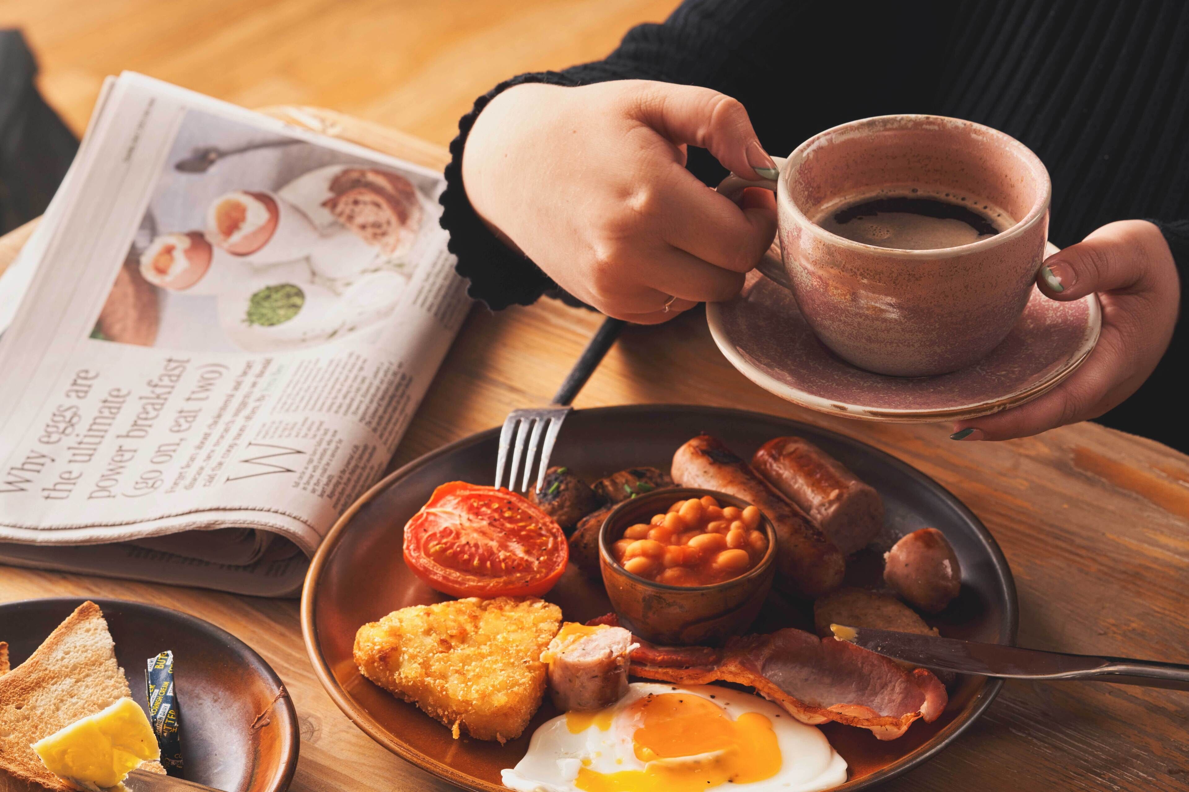 Prato de café da manhã com ovos, bacon, feijão, linguiça e torradas, uma xícara de café e um jornal sobre uma mesa de madeira.