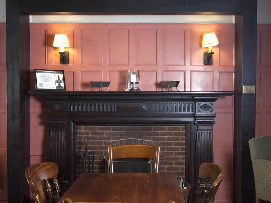 Uma sala de jantar com uma mesa e cadeiras de madeira, uma lareira de tijolos e duas luminárias fixadas na parede.