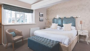 A bedroom with a large bed, a chair, a bench, a window with curtains, and a nightstand with a lamp. - The Globe (Exeter)