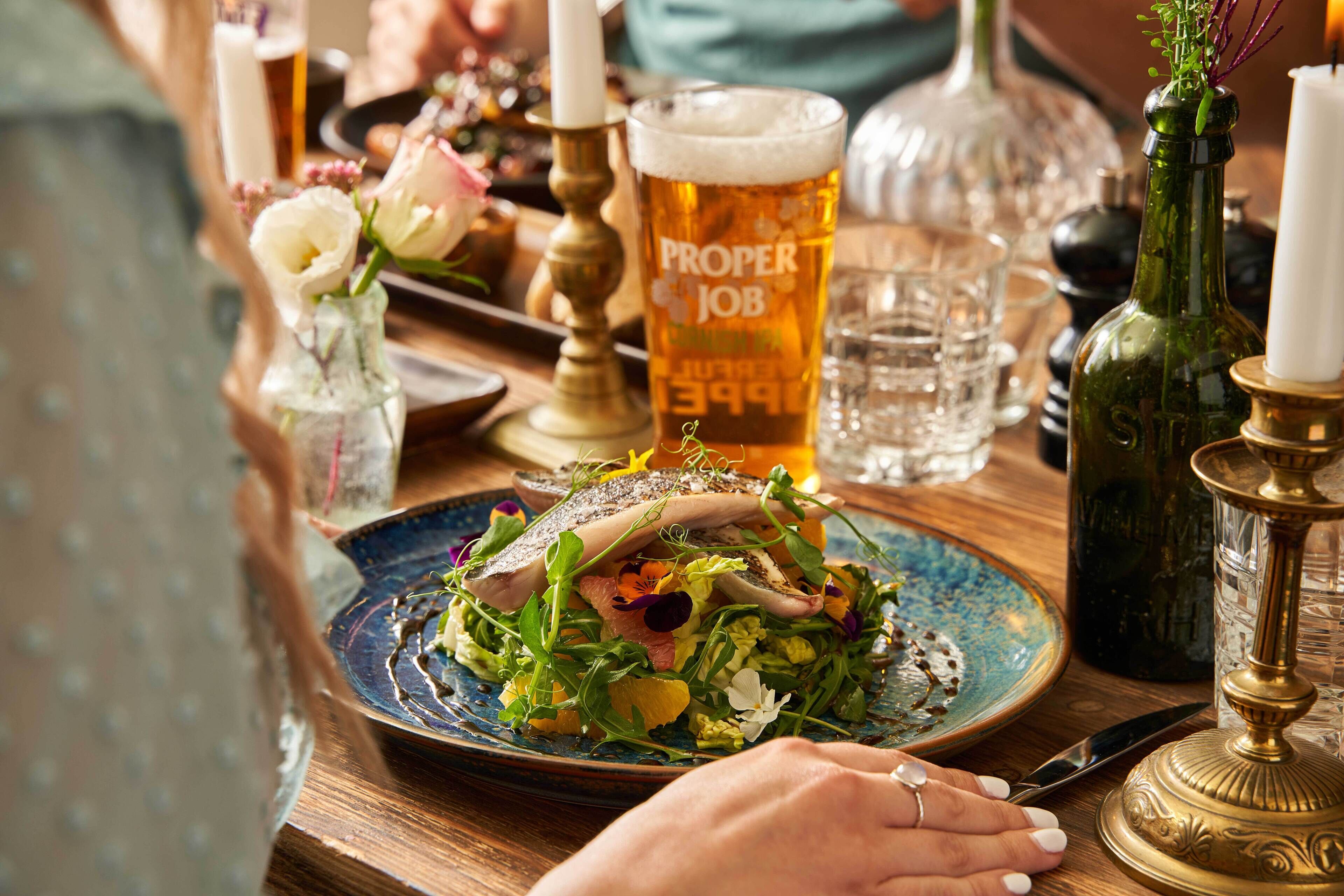 Una mesa de comedor con un plato de comida, una copa de cerveza con la etiqueta 'Proper Job' y varios utensilios de mesa.