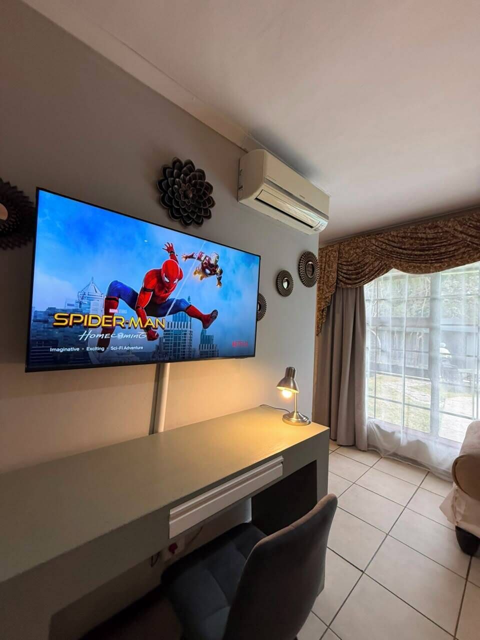 A living room with a flat-screen TV displaying a movie poster, a ceiling-mounted air conditioner, a window with curtains, and a sofa.