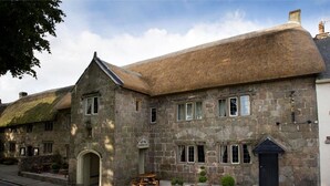 A stone building with a thatched roof, a wooden bench, and potted plants on the front.