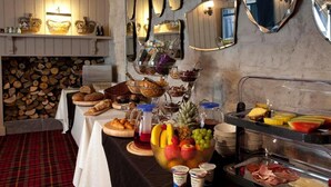 A buffet with various food items, including fruits, pastries, and beverages, arranged on tables with a plaid carpet and a stone wall in the background.